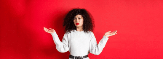 A imagem mostra uma mulher em um fundo vermelho vibrante. Ela tem cabelo cacheado e usa uma camiseta cinza simples. Sua expressão facial transmite dúvida ou incerteza, e ela está com as mãos levantadas e abertas, como se estivesse perguntando ou sem saber o que fazer. A pose e o olhar refletem uma sensação de questionamento ou confusão.