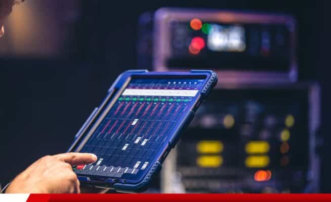 Mão operando mesa de som digital em tablet com equipamentos de áudio ao fundo.