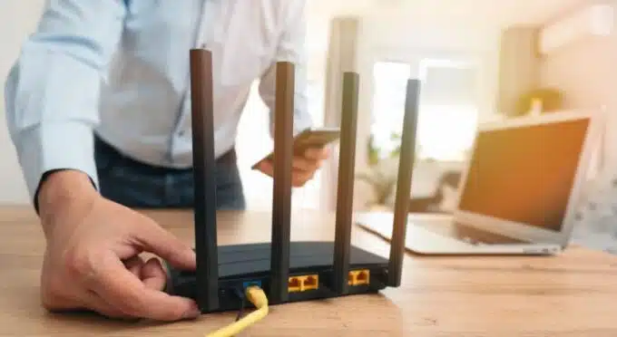 um roteador de internet sob uma mesa com um notebook, e um rapaz em pé tocando no roteador