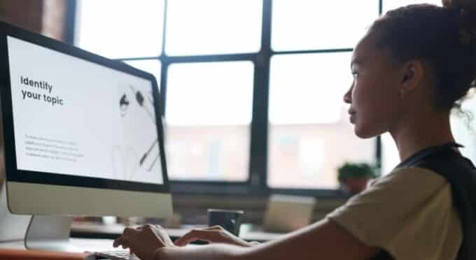 Uma jovem, com a pele escura e cabelo cacheado preso, é vista de perfil sentada em frente a um computador em um ambiente de escritório.