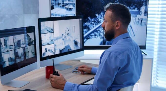 Um homem com barba, vestindo uma camisa social azul, está sentado de perfil em uma mesa, olhando atentamente para múltiplos monitores que exibem imagens de câmeras de segurança de um prédio residencial