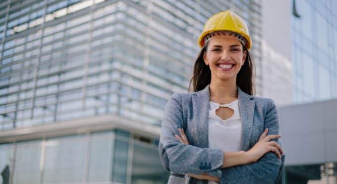 Uma mulher engenheira ou arquiteta sorridente, de cabelo escuro, usando um capacete de segurança amarelo. Ela veste um blazer cinza sobre uma blusa branca e está com os braços cruzados.