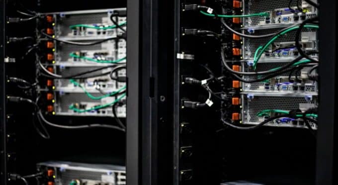 Servidores de computador montados em rack dentro de um gabinete ou centro de dados. Múltiplos cabos (incluindo cabos de rede pretos e cabos de alimentação verdes) estão conectados à parte traseira dos servidores