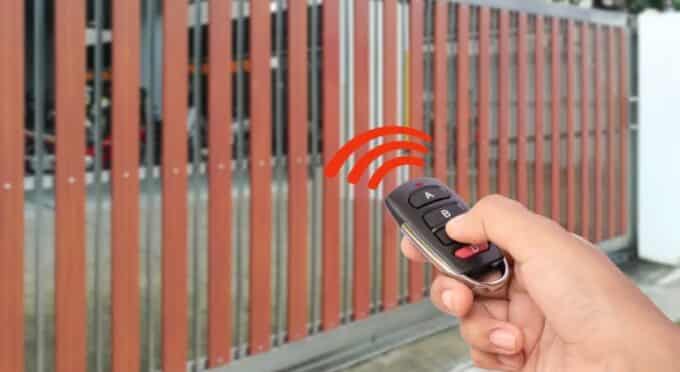 Uma mão segura um controle remoto preto com botão vermelho abaixo. Sinais de transmissão vermelhos em formato de ondas emanam do controle em direção a um portão de correr automático